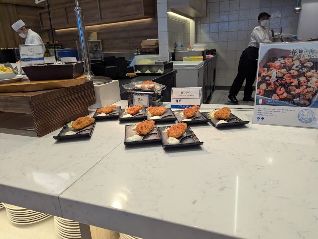鯵フライの小分け皿が並ぶコーナーをやや広角で撮った写真、奥にシェフと春の地中海フェアPOP
