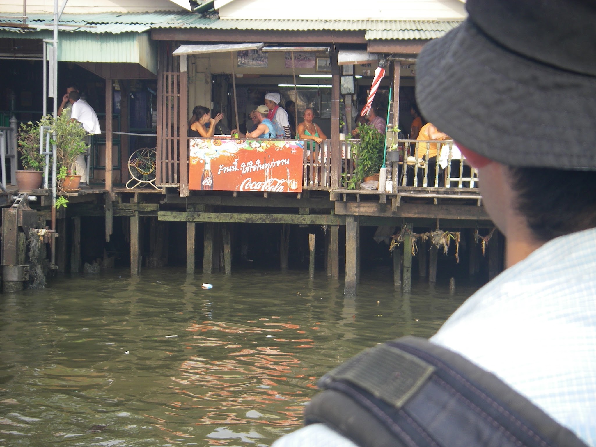コーラの看板を掲げた川沿いのテラスと、手前に写る同乗者の背中